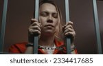 Small photo of Female prisoner in orange uniform holds metal bars, stands in jail cell in handcuffs. Woman criminal serves imprisonment term for crime in prison. Depressed inmate in detention center. Close up.