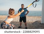 Small photo of Young personal trainer teaching how to do squats and leg strengthening exercises for her suspension training plan on the beach to an older white-haired person who huffs and puffs and attends.