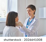 Friendly middle-aged woman doctor wearing gloves checking sore throat or thyroid glands, touching neck of young African female patient visiting clinic office. Thyroid cancer prevention concept