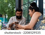 Small photo of A cheerful Indian woman, with dark curly hair, shares a moment of joy and laughter with her male partner, eagerly showing him her phone as they enjoy tea together at a coffee shop during brunch.