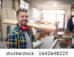 Portrait of professional carpenter holding wood plank material on his shoulder and smiling. Carpentry woodworking workshop in background.
