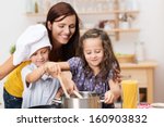 Small brother and sister cooking a meal both stirring the contents of the same pot watched over by their laughing young mother