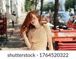 Small photo of Young redhead woman pouting at the camera with a thoughtful expression as she twiddles her long hair outdoors in town