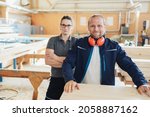 Small photo of Smiling carpenter wearing red ear muffs posing with a female colleague at a workbench in a large industrial woodworking factory