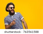 Young bearded hipster in cap and sunglasses pointing happily away on orange background. 