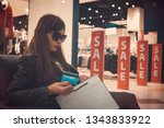 Small photo of Woman in dark glasses sitting in mall walking zone holding back of credit card and white paper shopping bag at sale signs background. Lady overspent during sale time and became upset. Use of credit