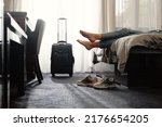 Small photo of woman taking off footwear in a hotel room on the bed. Tourist relaxing on hotel room after travelling with suitcase. Female having rest after long trip with language Dark silhouette