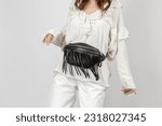 White Woman Wearing White Jeans, Blouse with Frills and Crossbody Black Leather Belt Bag with Decorative Fringe over Grey Background