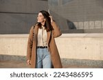 American shot of city girl touching her hair, wearing brown cloth coat, jeans, cream wool sweater, black shoulder-length straight hair, very white skin and wide smile. Background of gray buildings