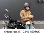 Stylish confident man, biker wearing helmet using mobile phone standing near sport motorcycle. Handsome fashion model wearing stylish leather jacket posing for pictures on motorbike 