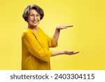 Smiling attractive senior woman holding hands with empty space looking at camera isolated on yellow background. Shopping, store, advertisement concept 