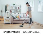 asian lady doing house chores in apron. young housewife using vacuum cleaner cleaning the wooden floor in the living room. happy housekeeper doing housework at home with attractive smile on face.