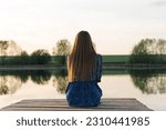 young beautiful caucasian woman in blue dress sits at wooden pier on river bank in summer at sunset, green trees and blue sky in background, melancholy concept, traveling along in vacation