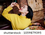 Funny brunette girl in yellow sweater eating pizza at restaurant. 