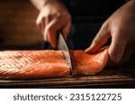 Chief hands cut salmon fillet with knife on wooden table at kitchen. Man cooking red omega fish with lemons for healthy nutrition diet