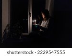 Small photo of Blackout. Energy crisis. Destruction of infrastructure. Power outage concept. Girl with a burning candle in a dark room sits near the window