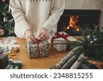 Woman wrapping stylish christmas gift with red ribbon on wooden table with festive decorations against fireplace in decorated scandinavian room. Merry Christmas! Hand holding present