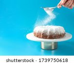 Small photo of Woman's hand sprinkling icing sugar over fresh home made bundt cake. Powder sugar falls on fresh perfect bunt cake over blue background. Copy space for text. Ideas and recipes for breakfast or dessert