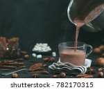 Pouring tasty hot chocolate cocoa drink into glass mug with ingredients on black table. Copy space Dark background. Low key.
