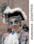 Small photo of Venice, Italy - 02.19.2022: A middle-aged man in a red vintage costume with a lace collar, wearing a carnival mask, tricorn hat with feathers stands at the carnival in Venice, Italy