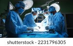 Small photo of Team of professional mixed-races doctors and assistants in medical uniforms performs heart transplant operation under lamp using medical tools in operating room looking at monitor. Surgery concept