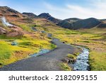 Pathway by stream flowing in mountain. Beautiful view of amidst green moss covered landscape against blue sky. Idyllic picturesque of volcanic valley in Alpine region.