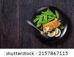Baked Cod with Crackers butter toppings served with boiled snow peas and lemon on black plate on dark wooden table, horizontal view from above, flat lay, free space, close-up