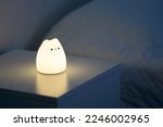 Cute cat shaped night lamp standing on a bedside table next to bed. Bedside lamp. Night lamp standing next to bed. Bedroom lamp on a night table next to a sleeping bed in a dark room. Close up. 