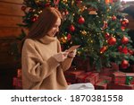 Side view of charming young woman getting Christmas greeting card from her beloved on background of xmas tree at cozy living room with festive interior. Surprised lady pleasure reading letter at home.