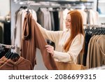 Side view of cheerful pretty young woman buyer choosing clothes from rack in clothing store, blurred background. Cute female shopaholic select and buying clothes in fashion boutique during sale.