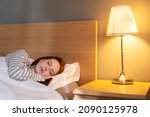 Small photo of Close-up face of tired young woman sleeping well in bed hugging soft white pillow at home, bedside lamp lighting with warm yellow light. Cute lady resting enjoying fresh soft bedding linen in bedroom