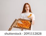 Small photo of Studio portrait of dissatisfied young woman eating bad quality pizza holding box in hands looking away standing on white isolated background. Pretty redhead female eating unappetizing meal.