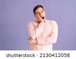 Portrait of exhausted and sleepy young handsome man yawning standing on purple isolated background in studio. Bored male feels sleepy, yawns as feels tired, opens mouth widely, closed eyes.