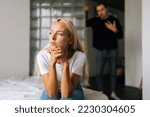 Small photo of Portrait of tired stressed woman sadness looking away sitting on bed on blurred background of aggressive husband shouting on wife at home. Concept of family scandal, crisis, domestic violence, abuse.