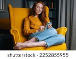 Portrait of happy redhead young woman using smartphone sitting in yellow armchair, smiling looking to device screen. Attractive ginger female holding phone in hands sitting in soft cozy chair at home.