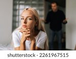 Portrait of tired young woman sadness looking at camera sitting on bed on background of aggressive husband shouting on girlfriend at home. Concept of family scandal, crisis, domestic violence, abuse.