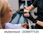 Small photo of Close-up cropped shot of unrecognizable orthodontist in rubber gloves showing artificial human jaws with dental braces for teeth correction to female patient, visiting doctor orthodontic issues.