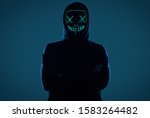 Portrait of an anonymous man in a black hoodie hiding his face behind a scary neon mask. Studio shot.