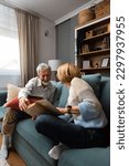 Small photo of Simple living. Happy middle-aged senior couple 70s husband and wife sit relax on couch in living room reading book drinking tea together, calm elderly 60s couple booklovers rest at home enjoy life