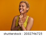 Pretty joyful African American with cute short curls, making faces in front of camera showing her tongue, after hitting wrist, having cheerful and carefree look, posing against orange wall in crop top