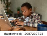 Small photo of Portrait of little African American boy in trendy shirt doing sneaky things, reaching his father's laptop, looking at camera with scared, canny face expression. Children misbehaving concept