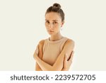 Small photo of Loneliness, body language. Pretty poor stylish girl of 20s in beige tank top standing against white studio background with sad upset facial expression, feeling pitiful, hugging herself