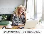 Attractive successful elderly businesswoman in striped blouse working in modern office, making phone call to potential client, having nice conversation, sitting at desk in front of open laptop