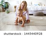 Cute cheerful European infant in diaper having joyful facial expression, laughing while crawling on floor from his smiling mother who is chasing him. Blonde young woman playing with son indoors