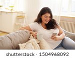 Beautiful cute young plus size female with loose dark hair enjoying cappuccino and surfing internet using mobile phone, watching videos via social networks, liking posts, leaving comments, smiling