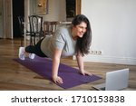 Sporty plus size woman in sportswear working out at home, doing plank on yoga mat in front of open laptop, repeating instructions by professional fitness trainer watching online video tutorial