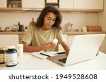 Small photo of Beautiful young female dietician working from home, using portable computer, typing article about vitamins, holding bottle of food supplements, reading label on it. Health, wellness and dieting