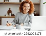 Small photo of Home portrait of pretty female English tutor working at home sitting at kitchen table, preparing for online lesson with her student, writing in her copybook in front of laptop and cup of coffee