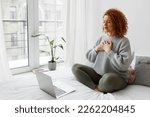 Small photo of Pretty redhead plus size female sitting on bed in lotus posture with hands on chest doing pranayama techniques in front of laptop, breathing deeply, reaching inner balance after stressful day