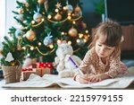 Cute little child girl writing letter to Santa Claus or writing dreams of a gift with near Christmas tree. Merry Christmas and Happy New Year!